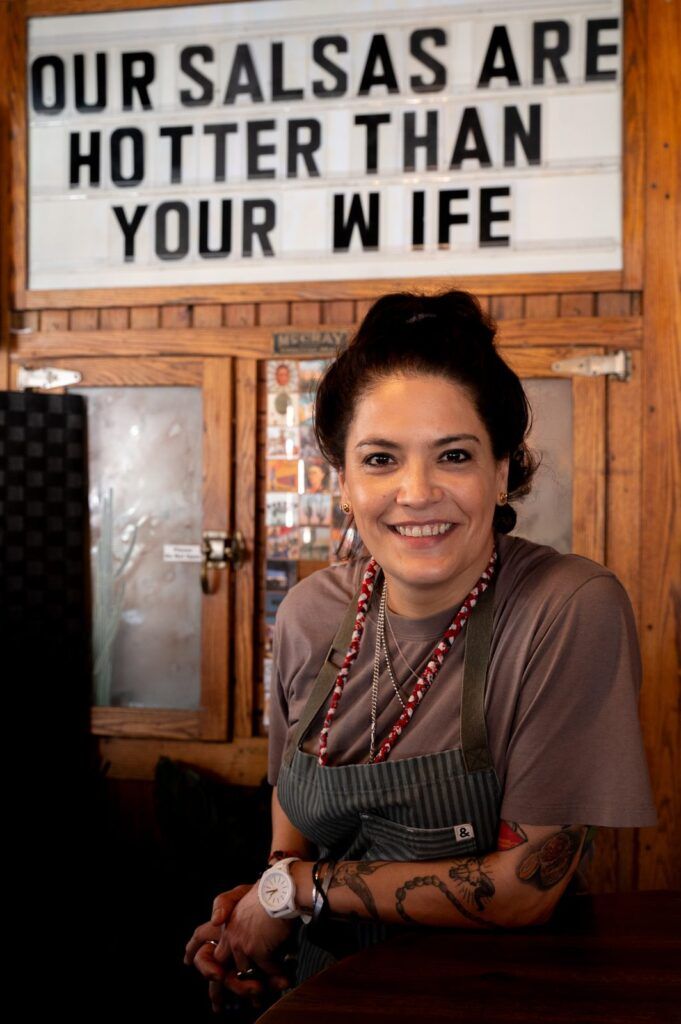 Chef MARIA MAZON de Tucson sonriendo frente a un letrero sobre salsas picantes, cocina con humor e identidad fronteriza