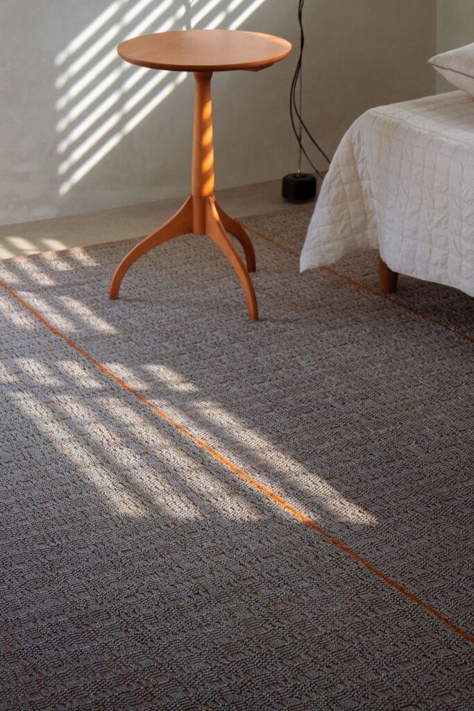 Alfombra de otoño Paglietta Cotta de G.T.DESIGN en yute gris con línea naranja en una recámara con mesa auxiliar de madera y luz natural