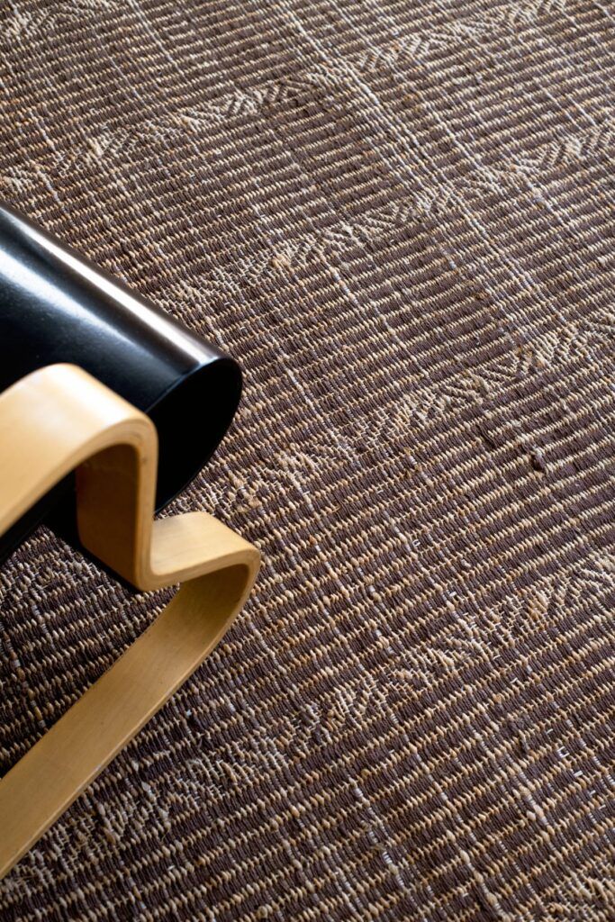 Detalle de la alfombra de otoño Sahil Terra de G.T.DESIGN en yute natural con motivos geométricos junto a un sillón de madera curvada